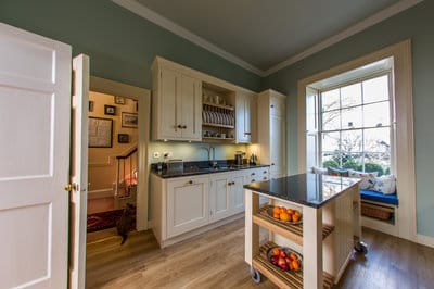 wide view of kitchen showing mobile kitchen island, window seat,run of cabinets with sink, drainage and crockery storage